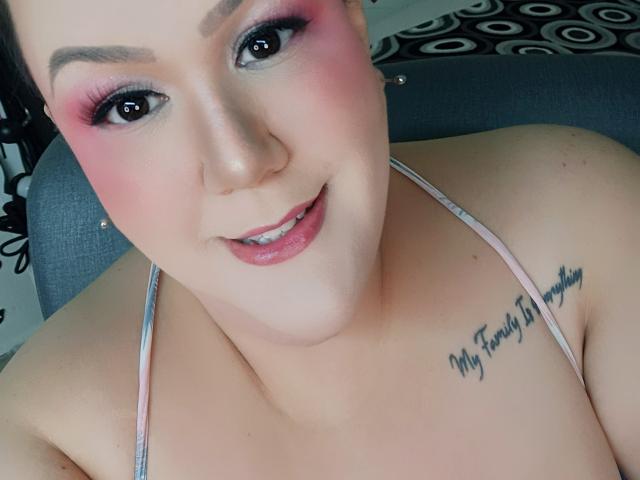 GiiannaMorrys with pink eyeshadow, glossy pink lips, and a tattoo on her right shoulder that reads "My family is everything." Her tits are visible, wearing a spaghetti strap top.