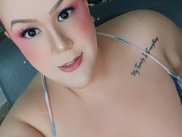GiiannaMorrys has pink eye makeup, glossy pink lips, and a tattoo on her right shoulder. She's topless with visible breasts, wearing a pink and blue halter top.