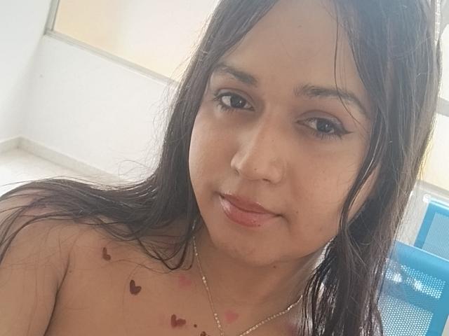 VivienSandrina heeft nat, donkerbruin haar, bruine ogen en kleine, rode hart-tatoeages op haar borst. Ze draagt een zilveren ketting en kijkt met een lichte glimlach naar de camera..