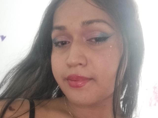 VivienSandrina heeft donker, steil haar, paarse oogschaduw, zwarte eyeliner, roze lippenstift en een lichte, ontspannen glimlach. Ze draagt een zwarte band over haar schouder, en haar huid is licht..