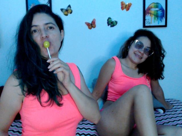 LunaVenus with dark hair, pink tank top, and yellow lollipop, sitting on bed, next to LunaVenus with curly hair, glasses, and pink tank top, smiling.
