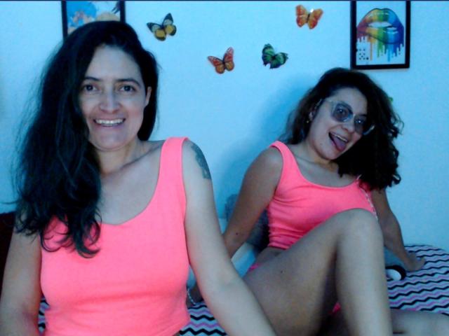 LunaVenus with dark hair, wearing a neon pink tank top, showing her tits, sitting on a bed with her ass up, smiling, and another LunaVenus with glasses in the background.
