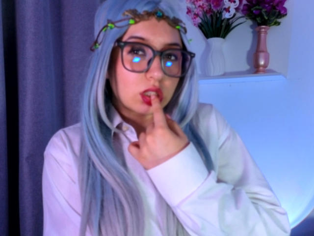 SusanMonroy with blue hair, glasses, and a flower headband, sucking on her finger, wearing a white shirt, in a slightly blurry photo, looking cute and sexy.