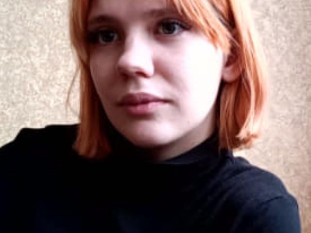 Louise Carsten, com a pele clara e um corte de cabelo curto laranja, veste uma gola alta preta, tem uma expressão neutra e lábios pequenos, ligeiramente entreabertos. Suas nádegas não estão visíveis..