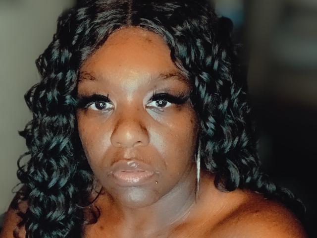 MzThundaBooty, con cabello rizado oscuro, piel morena, un pequeño piercing en la nariz y aros, luce seria con un maquillaje sutil y hombros descubiertos. Sus senos son parcialmente visibles..
