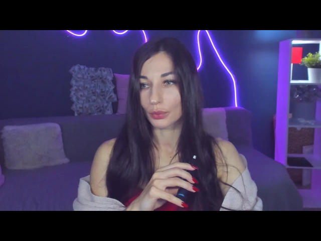 AlexOakman tem cabelos longos e escuros, batom vermelho, e está segurando uma garrafa com unhas vermelhas, vestindo uma blusa com um ombro descoberto, com um piercing nasal fofo, em um quarto com iluminação azul..