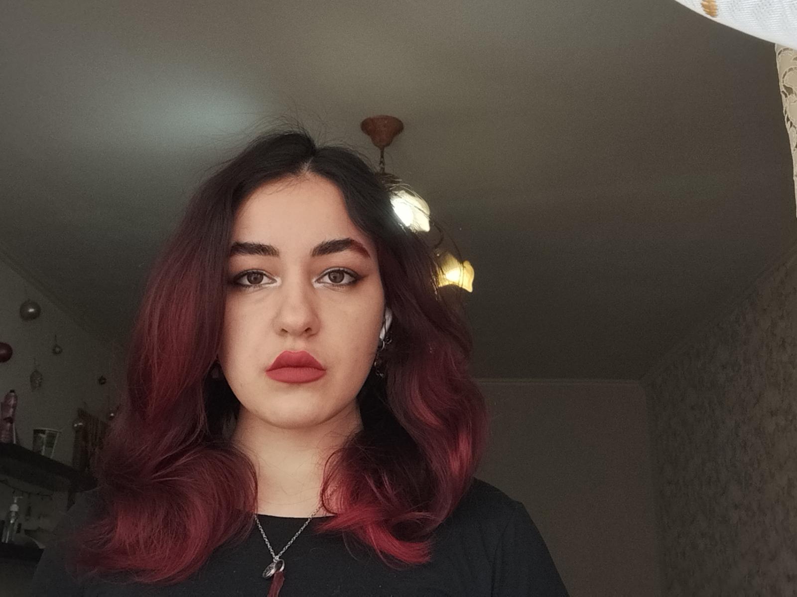 Melody Galindo ha i capelli con sfumature dal rosso al nero, labbra piene e un'espressione seria, indossa una maglietta nera con una collana di gemme rosse e auricolari bianchi..