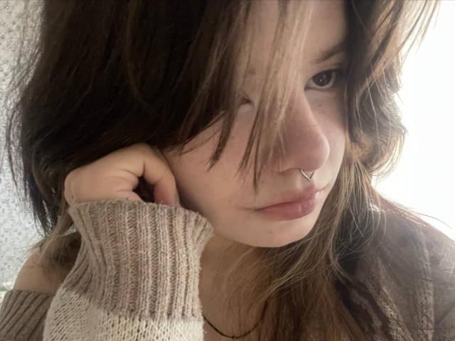 MinShutes with messy brown hair, nose ring, and pouty lips, wearing a beige knit sweater, touching her cheek. Soft light highlights her gentle expression.