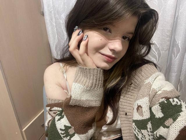 MinShutes with brown hair, wearing a camo cardigan, holding her cheek, showing her tits in a white bra, and having patterned nails, in a soft lit room.