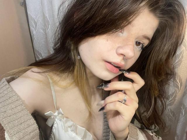 MinShutes has brown, wavy hair, a nose ring, and is biting her finger with black, glittery nails. She's wearing a beige cardigan and a white, ruffled top.