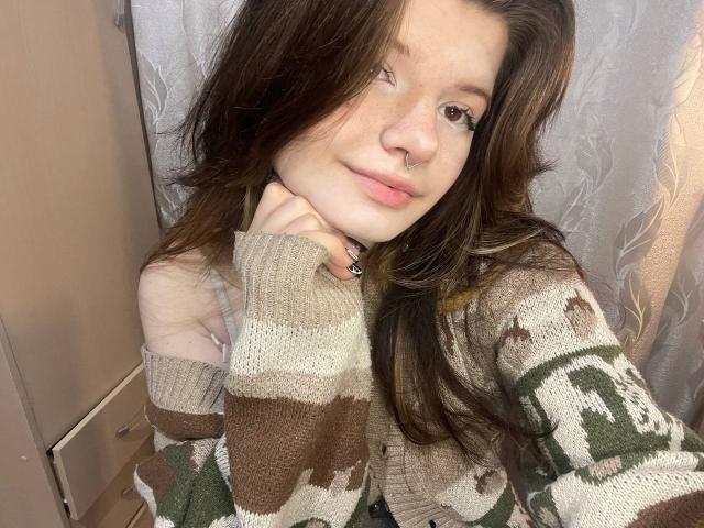 MinShutes with long brown hair, freckles, nose ring, and green/brown/white patterned sweater, resting chin on hand, smiling softly, wearing black ring.