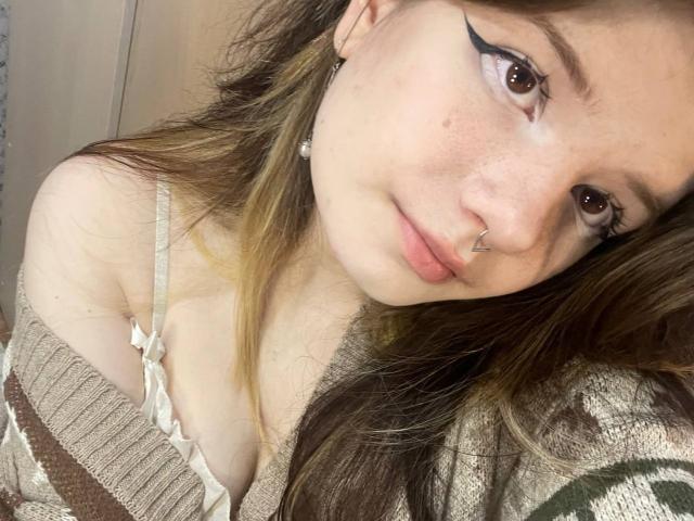 MinShutes with brown eyes, nose ring, and black eyeliner, wearing a beige off-shoulder top showing her tits, long brown hair, and earrings, looking at the camera.