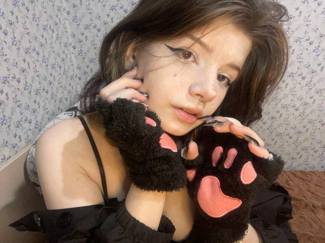 MinShutes with cat paw gloves, black dress, and pink paw pads, looking at camera, nose ring, black eyeliner, and brown hair, showing tits, sitting on brown bed.