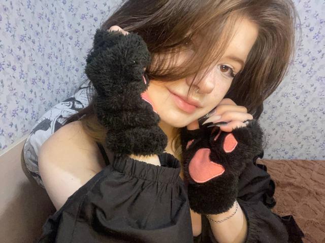MinShutes with brown hair, wearing black paw gloves with pink pads, holding a black stuffed cat, smiling softly, wearing a black off-shoulder top, in a cozy setting.