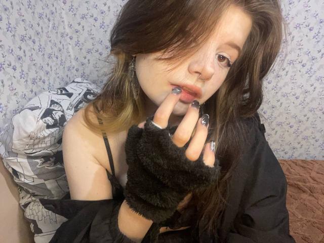 MinShutes with dirty blonde hair, black fingerless glove, and painted nails, biting her finger, showing her tits in a black top, sitting on a bed with a floral background.