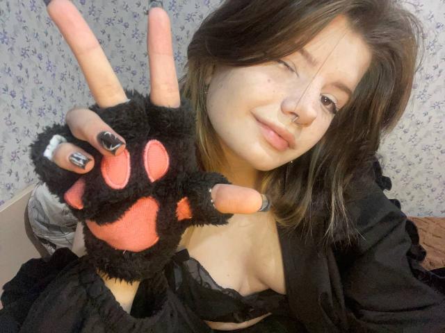 MinShutes with brown hair, nose ring, and black fingerless glove showing a peace sign, winking, wearing a black lace bra, and a black top, smiling. Boobs visible.