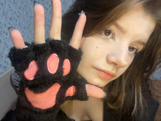 MinShutes with a cute black furry paw glove, pink pads, holding it up. She's got brown eyes, a nose ring, and a serious look. Her tits are partially visible.