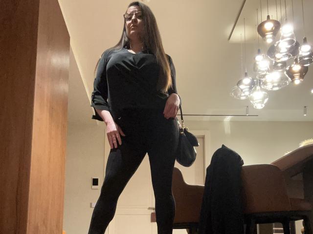 BadAngel with long brown hair, glasses, black shirt, black pants, holding black bag, standing in dimly lit room, looking down, hands on hips, wooden wall on left.