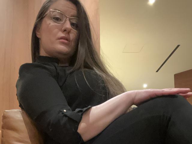 BadAngel with long brown hair, wearing glasses and black clothes, sitting with arms crossed, looking slightly annoyed. She has a small nose and is in a wooden room.