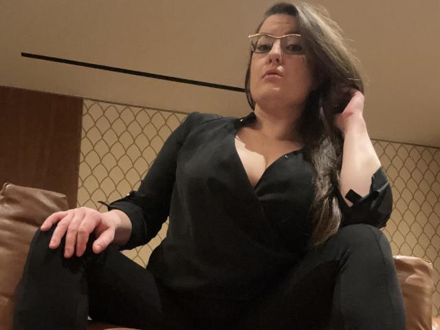 BadAngel, a chubby woman with glasses and long brown hair, sits on a couch, wearing a black shirt with deep cleavage and black pants, hand in hair, looking down.