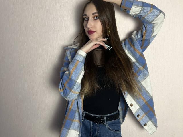IndiaClune with long brown hair, blue plaid shirt, black top, jeans, and belt, hand in hair, red lipstick, standing against pink wall, looking at camera.