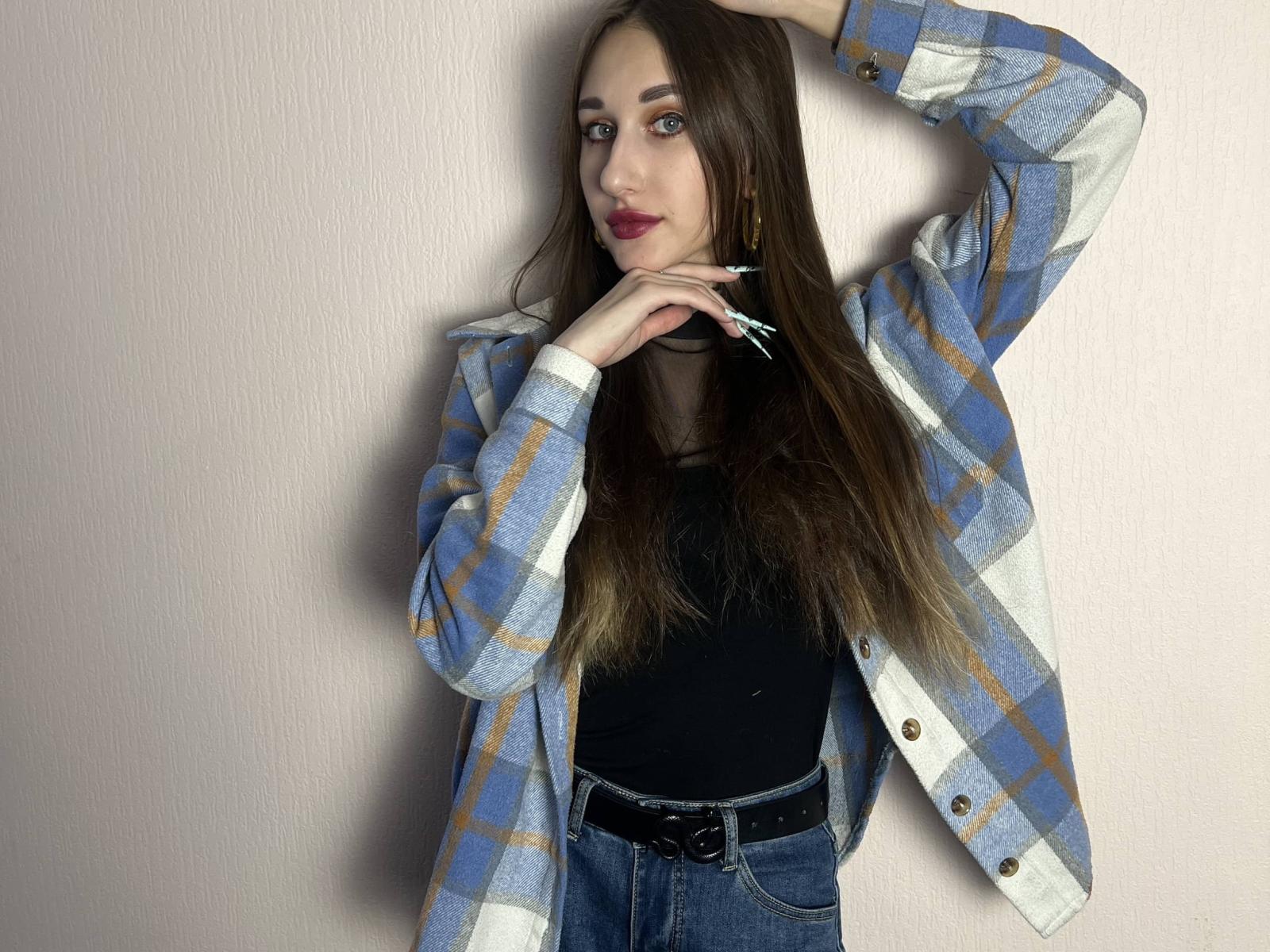 IndiaClune with long brown hair, blue plaid shirt, black top, jeans, and belt, hand in hair, red lipstick, standing against pink wall, looking at camera.