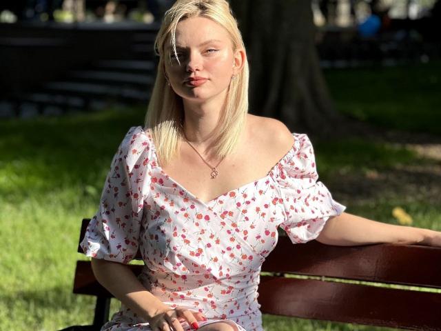 CrystalHope, con cabello rubio, sentada en un banco, vestida con un vestido blanco de corte asimétrico con estampados de fresas rosas, con la luz del sol en su rostro, una mano sobre su muslo, y un collar con un pequeño corazón..