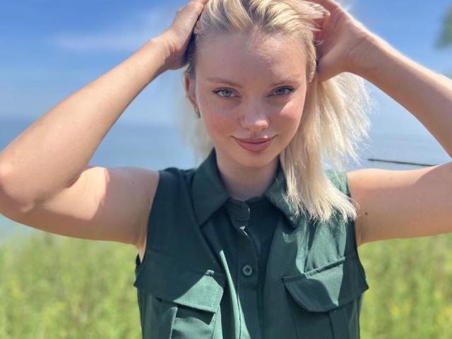 CrystalHope has blonde hair, blue eyes, and a cute smile. She's wearing a green sleeveless shirt, hands in her hair, with visible tits and a slim, attractive body.