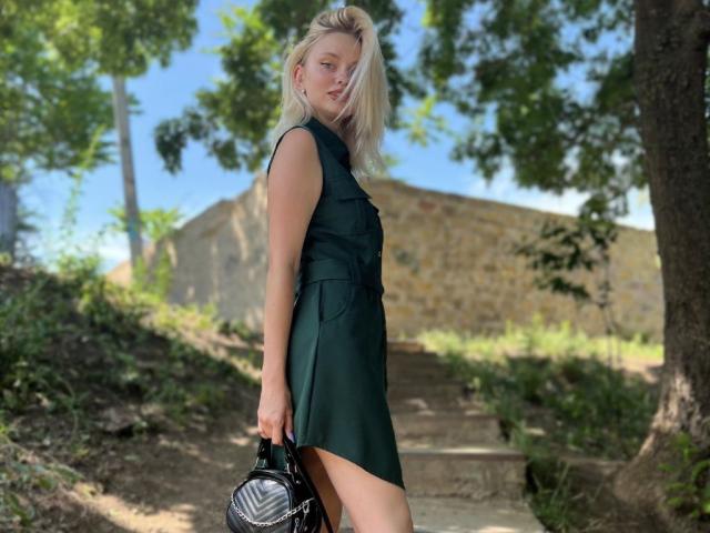 Blonde CrystalHope in a green dress, holding a black backpack, stands on sunlit stairs, her ass slightly exposed, with trees in the background. Her tits are small.