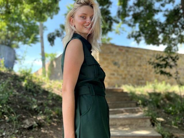 CrystalHope with blonde hair, smiling, wearing a sleeveless dark green dress, standing on stone steps, showing her ass, with trees and a stone wall in the background.