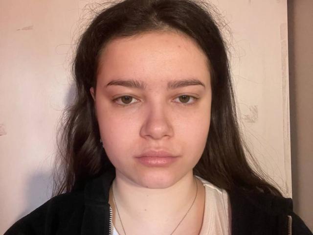 LashaunDromgoole heeft bleke huid, bruine ogen en lang donker haar, en draagt een zwarte jas over een wit shirt, met een neutrale, lichtjes fronsende uitdrukking op haar gezicht..