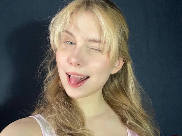 ShenitaMarts has blonde, wavy hair, is winking with her right eye, and has her tongue slightly out, showing a playful expression. She's wearing a white tank top.