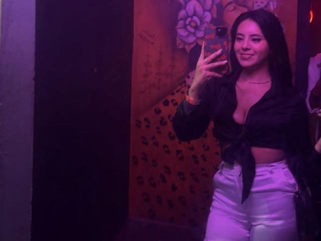 EimmyPark with long dark hair, smiling, wearing a tied-up black top showing her tits and white pants, taking a mirror selfie with a pink phone.