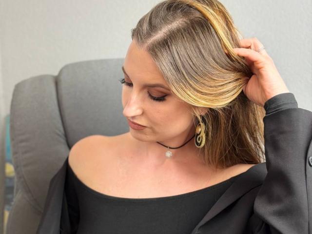 Milagres with blonde and brown hair, eyes closed, black off-shoulder top, black jacket, and gold earrings, holding hair, wearing a black choker with a small pendant.