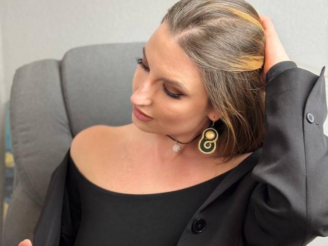 Milagres with her brown hair, partially blonde, in a black off-shoulder top, wearing a black buttoned jacket, and a black and gold swirl earring, touching her head.