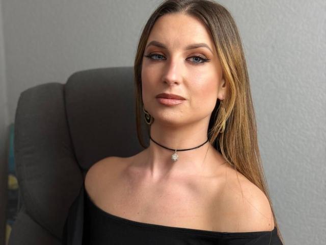Milagres with long brown hair, blue eyes, and off-shoulder black top, wearing a black choker with a small pendant, sitting in a gray chair, looking straight at camera.