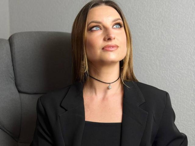Milagres, with blue eyes and straight brown hair, wears a black blazer over a black top, a black choker with a small pendant, and sits in a grey chair.
