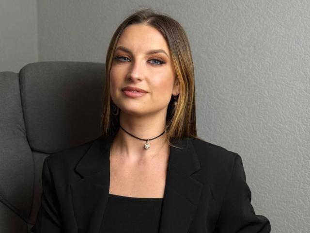 Milagres has straight brown hair, blue eyes, and wears a black blazer with a black choker. Her tits are small, and she's sitting in a gray chair.