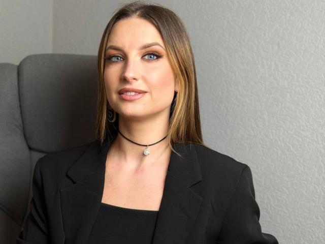 Milagres has straight brown hair, blue eyes, and a subtle smile. She's wearing a black blazer, black top, and a black choker with a small pendant.