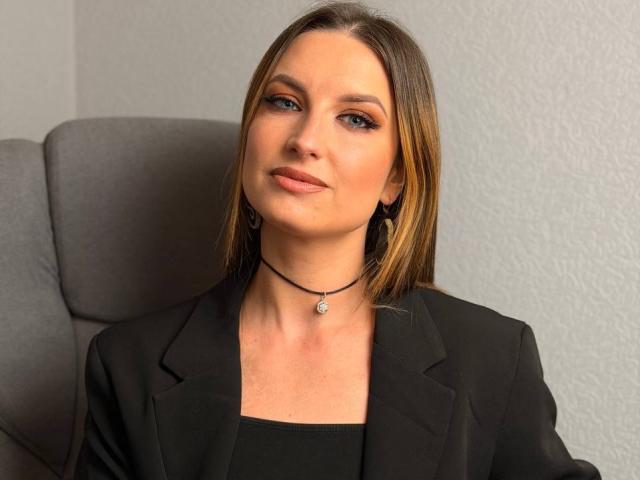Milagres has straight brown hair, subtle makeup, and a black blazer. She's wearing a black choker with a small pendant, sitting in a gray office chair.