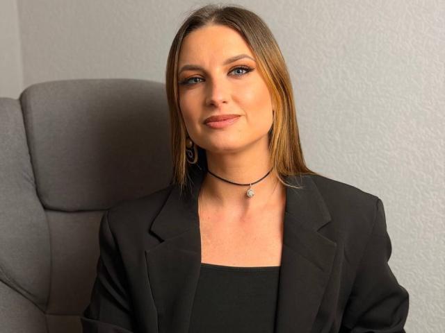 Milagres has straight brown hair, blue eyes, and is wearing a black blazer over a black top, with a black choker and small earrings, sitting in a gray chair.