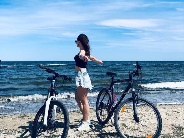 Milagres with tits in a black top, denim shorts, and white socks stands on a beach, arms out, between two bikes with a blue ocean and white sky.