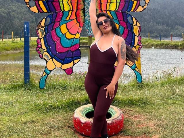 MelizaQueen with long brown hair, wearing a tight maroon one-piece, standing in front of a colorful butterfly mosaic, arm raised, tattoo on left arm, wearing sunglasses.