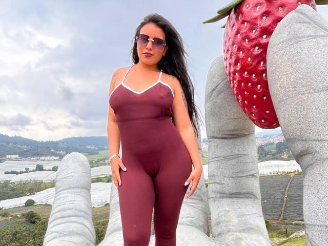 MelizaQueen with dark hair, sunglasses, and a tight maroon one-piece, showing her ass and pussy through the fabric. Large strawberry statue in the background.