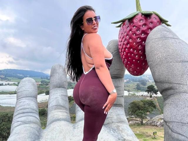 MelizaQueen with long black hair, wearing sunglasses and a maroon one-piece, showing her ass, standing in front of a giant gray hand holding a purple strawberry.
