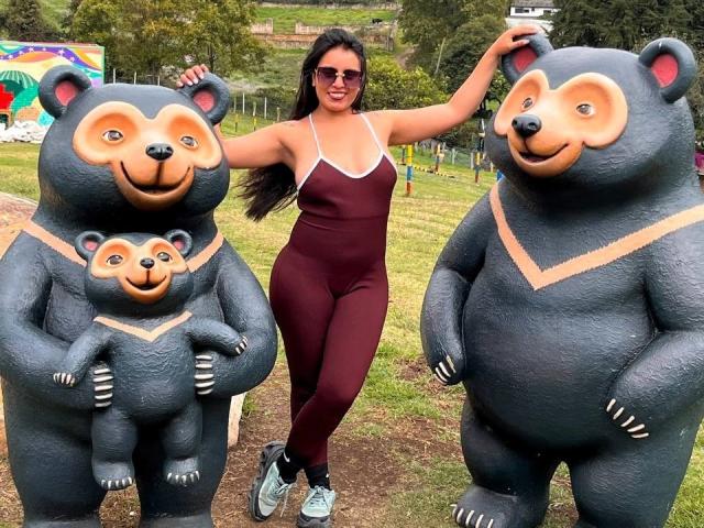 MelizaQueen with big ass in maroon one-piece, standing between two giant, cartoon bear statues, one holding a small bear, wearing sunglasses, in a grassy park.