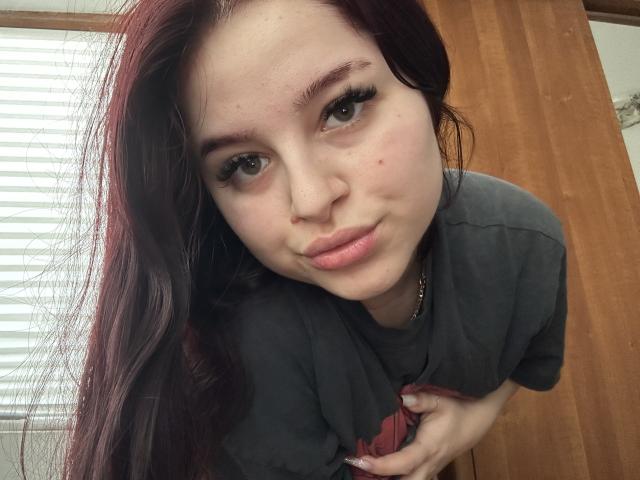 LucieRed with long dark red hair, wearing a gray shirt, holding her tits, looking up with a slight smile, brown eyes, subtle makeup, and a small zit on her nose.