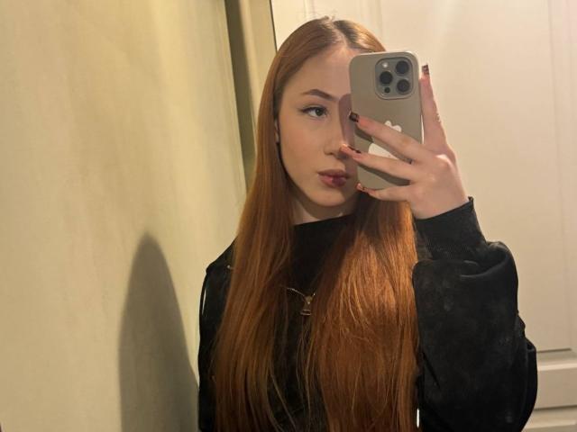 AlonaHadcock with long red hair, black sweater, holding a phone in front of her face, looking to the side, in a mirror selfie. Boobs are not visible.