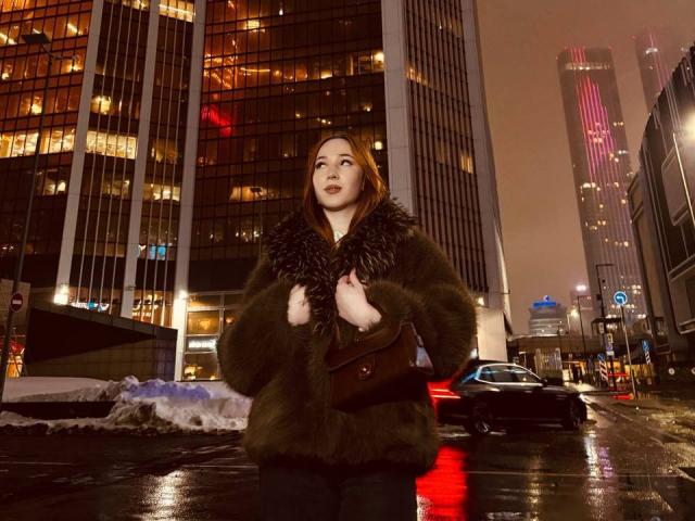 AlonaHadcock with red hair, wearing a dark fur coat, holding a brown purse, standing on a wet, reflective city street at night, with tall buildings and neon lights.