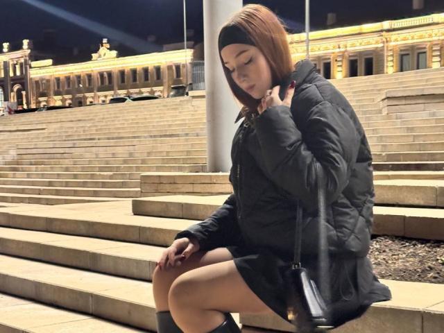AlonaHadcock, with short brown hair and a black headband, sits on stone steps, wearing a black puffy jacket, black skirt, and black boots, holding a black purse.